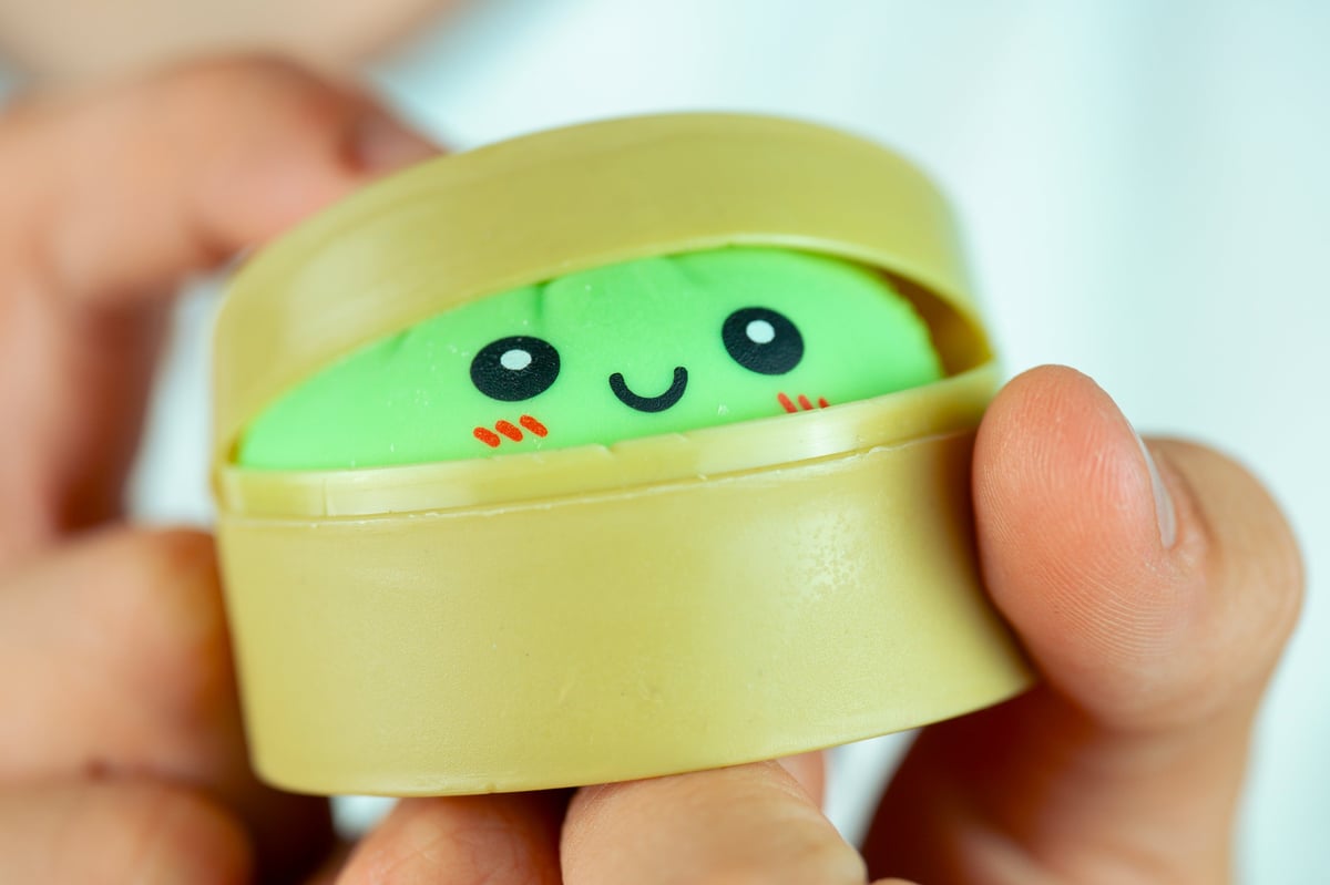 A hand holding a cute green kawaii dumpling squishy toy inside a steamer container